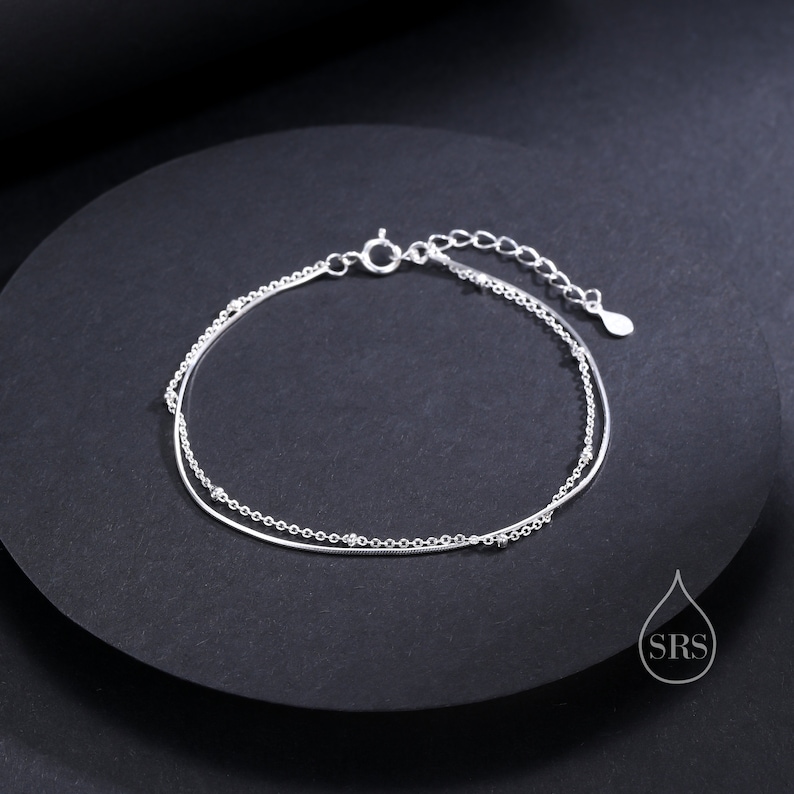 May include: A double-strand silver bracelet featuring one smooth snake chain and one delicate cable chain accented with small, evenly spaced silver beads. The bracelet includes a spring ring clasp and an adjustable extender chain with a teardrop charm.