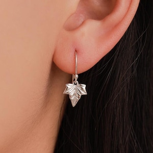 May include: A silver-colored earring with a leaf design. The earring is a small, detailed representation of a leaf, with visible veins and a pointed tip. The earring hangs from a simple hook.
