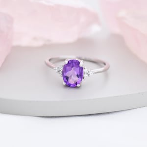 Könnte beinhalten: Ein silberner Ring mit einem großen, oval geschliffenen, lila Amethyst-Edelstein. Der Ring hat kleine, klare Akzentsteine auf beiden Seiten des Hauptsteins. Der Ring liegt auf einer hellgrauen Oberfläche mit einem zartrosa Hintergrund.