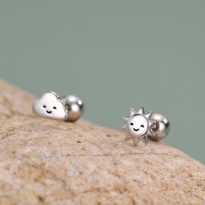 May include: Silver-colored stud earrings featuring a smiling cloud and a smiling sun design. The cloud earring is a puffy shape, and the sun earring has spiky rays. Both earrings have a small, round backing.