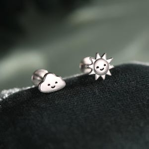 May include: A pair of silver stud earrings featuring a cloud and a sun design. The cloud earring has a smiling face, and the sun earring has a smiling face with pointed rays. The earrings are small and delicate.