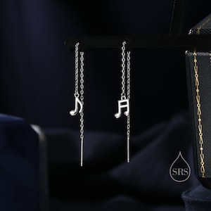 Peut inclure: Une paire de boucles d'oreilles pendantes en argent avec un charme en forme de note de musique. Les boucles d'oreilles sont sur un fond noir.