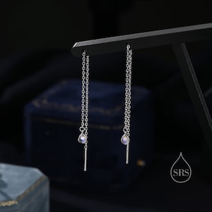 May include: A pair of silver dangle earrings with a teardrop-shaped moonstone pendant on each earring. The earrings are hanging from a black display stand.