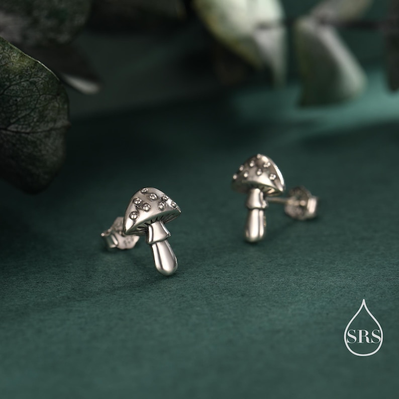 May include: A pair of silver mushroom stud earrings. The mushroom caps are dotted with small, clear stones. The earrings are set against a dark green background, with a blurred leaf in the upper left corner.
