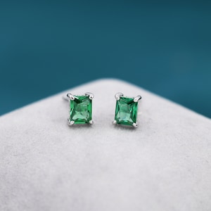 May include: A pair of square-cut, emerald-green gemstone stud earrings. The gemstones are set in a silver-coloured metal with four prongs. The earrings are displayed on a light grey surface against a teal background.