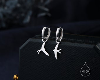 May include: A pair of silver hoop earrings with a dangling swallow charm. The earrings are on a black background.