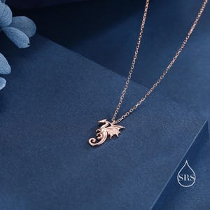 May include: A rose gold-toned necklace featuring a delicate dragon-shaped pendant suspended from a fine link chain. The dragon is depicted in a curled, stylized pose with visible wings and a tail. A small logo with the letters SRS is visible.