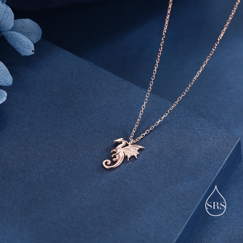 May include: A rose gold-toned necklace featuring a delicate dragon-shaped pendant suspended from a fine link chain. The dragon is depicted in a curled, stylized pose with visible wings and a tail. A small logo with the letters SRS is visible.
