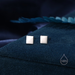 May include: A pair of silver square stud earrings on a blue velvet background.
