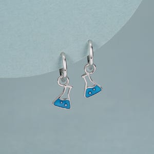 Könnte beinhalten: Silberne Creolen-Ohrringe mit blau-weiß emaillierten Kolbenanhängern. Die Kolbenanhänger haben einen abgerundeten Boden und einen schmalen Hals, mit drei kleinen weißen Punkten im blauen Emaille. Die Ohrringe sind vor einem hellblauen Hintergrund abgebildet.