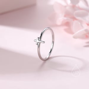 以下が含まれることがあります： シンプルな十字架のデザインのシルバーリング。リングは細くて繊細で、十字架はバンドの中央に配置されています。