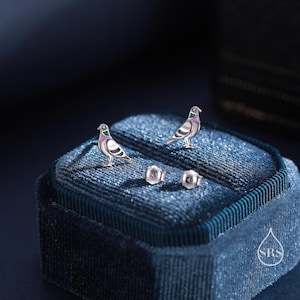 May include: A pair of silver dove earrings with green and pink accents, displayed in a blue velvet jewelry box with the logo "SRS".