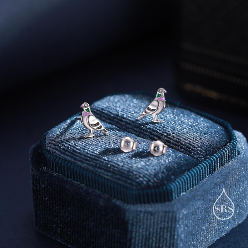 May include: A pair of silver dove earrings with green and pink accents, displayed in a blue velvet jewelry box with the logo "SRS".