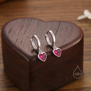 May include: Silver hoop earrings with heart-shaped, ruby-red pendants. The earrings are presented on a dark brown, heart-shaped wooden box. The earrings have a polished finish and a secure clasp.