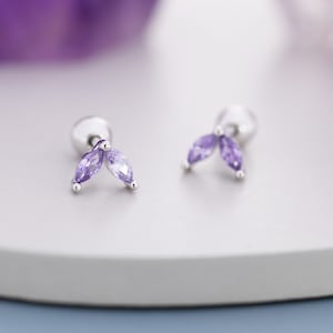 Puede incluir: Un par de pendientes de plata con diseño de hoja. Cada pendiente tiene dos piedras preciosas de color púrpura claro con corte marquesa y una pequeña bola plateada. Los pendientes se muestran sobre una superficie gris claro con un fondo púrpura borroso.