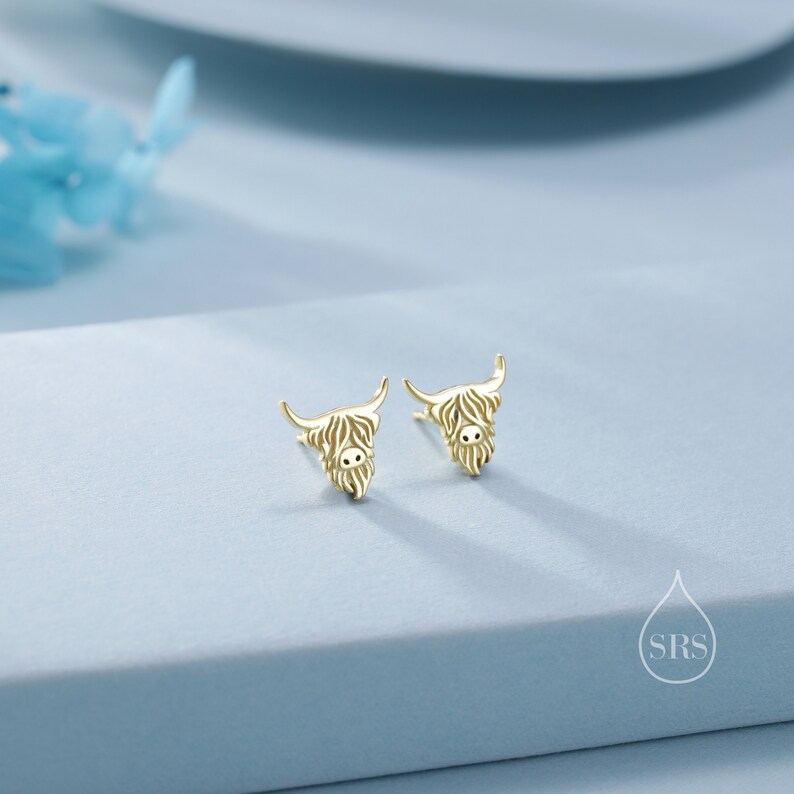 Puede incluir: Un par de pendientes de bot&oacute;n dorados con forma de cabeza de vaca Highland. Cada pendiente presenta un dise&ntilde;o texturizado detallado que representa el pelo largo y desgre&ntilde;ado del animal, cuernos curvados y una peque&ntilde;a nariz, con una superficie pulida.