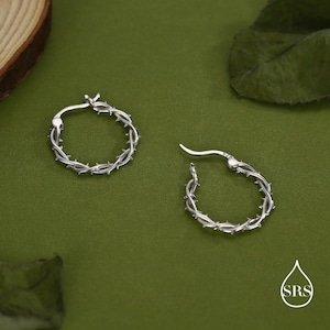 May include: A pair of silver-colored hoop earrings. Each earring is designed to resemble a thorny vine, with small, pointed thorns encircling the hoop. The earrings are displayed against a green background.