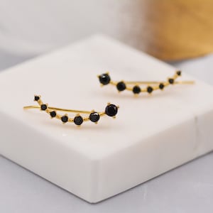 May include: A pair of gold ear cuffs with black gemstones. The cuffs are curved and have a minimalist design.