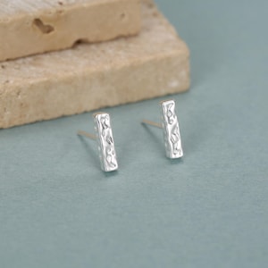 May include: A pair of silver bar stud earrings with a hammered texture. The earrings are rectangular and have a textured surface. The earrings are displayed on a light blue surface with a beige stone in the background.