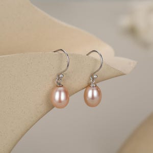 May include: A pair of drop earrings with peach-coloured, oval pearls. The pearls hang from delicate, silver-toned fish hook ear wires. The earrings are presented against a neutral background, emphasising their refined design. The pearls are a soft, muted shade.