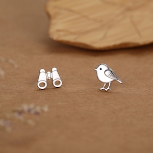 Puede incluir: Pendientes de plata con forma de pájaro y prismáticos. El pájaro tiene marcas detalladas en las alas. Los prismáticos tienen dos lentes circulares. Los pendientes están sobre un fondo marrón cálido.