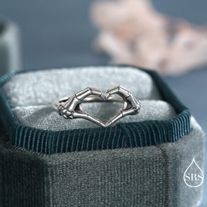 May include: A silver-toned ring featuring two skeletal hands positioned to form a heart shape. The metal has a polished finish with dark recessed detailing to highlight the bone structure of the fingers and wrists. The ring is centered in the frame.