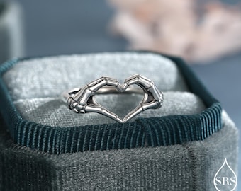 Puede incluir: Un anillo de tono plateado que presenta dos manos esqueléticas posicionadas para formar una figura de corazón. El metal tiene un acabado pulido con detalles oscuros en relieve para resaltar la estructura ósea de los dedos y las muñecas.