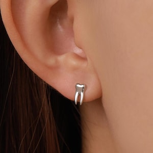 May include: A small, polished silver-tone stud earring shaped like a human tooth with two distinct roots. The earring features a smooth, reflective metallic surface and is secured directly into the earlobe, showcasing a simple and clean design.