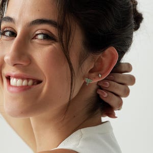 May include: A pair of stud earrings featuring a small, light green moth with dark outlines and a tiny, clear, round gemstone stud positioned above it, both worn on the earlobe to create a delicate and detailed insect-themed jewelry set.