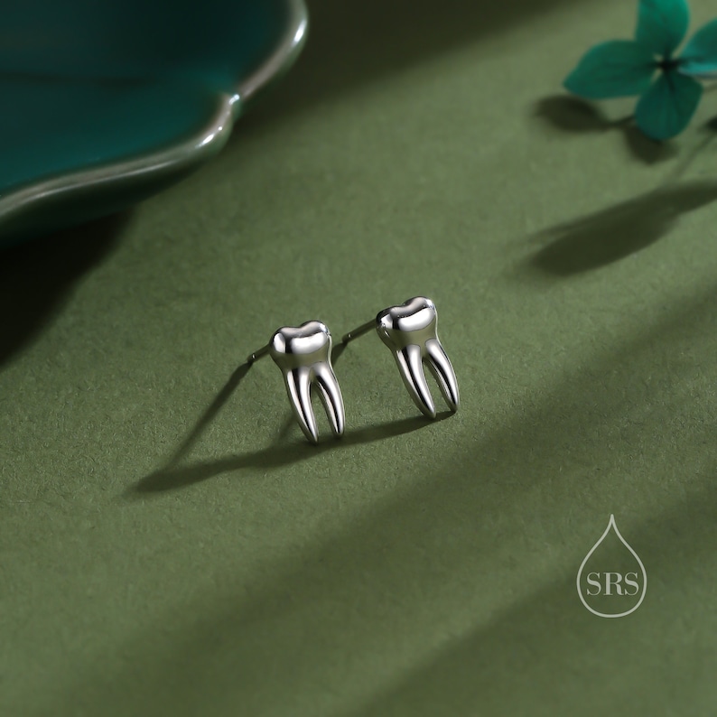 May include: A pair of silver-toned stud earrings shaped like human molars with two distinct roots. The earrings feature a smooth, polished metallic finish and are designed with a standard post backing for secure attachment to the earlobe.