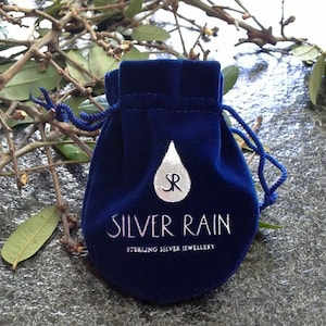 以下が含まれることがあります： 白い紐が付いた青いベルベット製のポーチ。ポーチには「SILVER RAIN STERLING SILVER JEWELLERY」という白いロゴと、白い涙の形をした「SR」の文字が入っています。