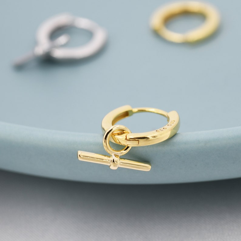 May include: Gold hoop earrings with a small gold bar charm hanging from the hoop.