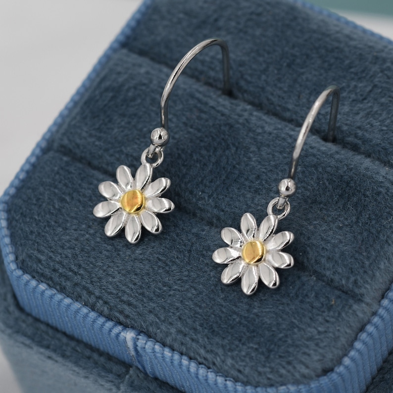 May include: A pair of silver daisy earrings with gold centers. The earrings have a hook design and are displayed on a blue velvet surface. The daisy petals are silver, and the center is gold.