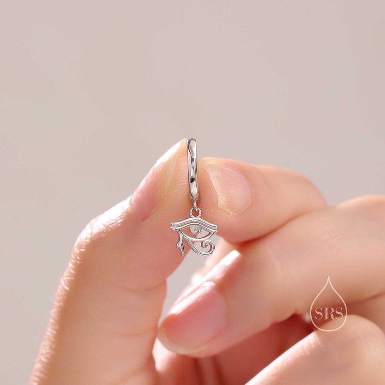 May include: Silver hoop earring with a dangling charm featuring a detailed silver eye of Horus design with a small clear stone in the center.