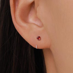 Puede incluir: Primer plano de una oreja con un pequeño pendiente de botón de piedra preciosa roja y redonda. El pendiente está engastado en metal plateado. También es visible un pequeño pendiente de aro plateado.