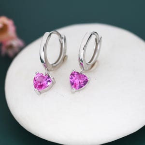 May include: A pair of silver hoop earrings with heart-shaped, bright pink gemstones. The earrings are displayed on a white, rounded surface, with a dark green background. The gemstones are a vibrant pink color.