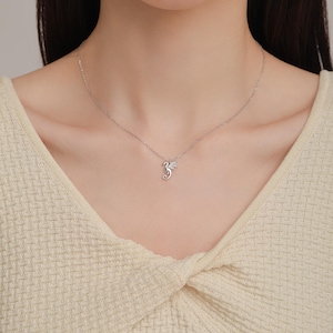 May include: A delicate silver-toned necklace featuring a small, stylized dragon pendant. The pendant is suspended from a thin, fine-link chain that rests against the collarbone. The dragon design is compact with visible wing and tail details.