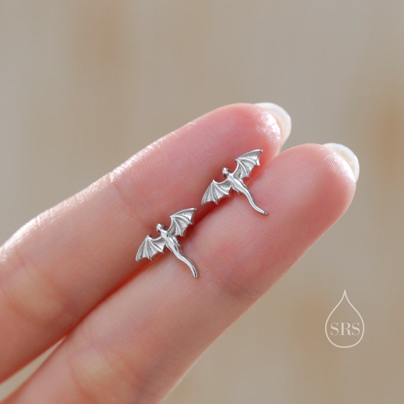 K&ouml;nnte beinhalten: Ein Paar silberne Drachen-Ohrstecker. Jeder Ohrring zeigt einen detaillierten Drachen mit ausgebreiteten Fl&uuml;geln und einem gebogenen Schwanz. Die Ohrringe sind klein und filigran.