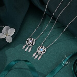 May include: Two silver-toned necklaces featuring circular dreamcatcher pendants with intricate web patterns and three dangling leaf-shaped charms. Each pendant is centered with a small, round, iridescent stone. A small logo with the letters SRS is visible.