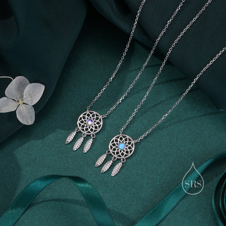 May include: Two silver-toned necklaces featuring circular dreamcatcher pendants with intricate web patterns and three dangling leaf-shaped charms. Each pendant is centered with a small, round, iridescent stone. A small logo with the letters SRS is visible.