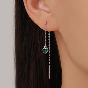 Könnte beinhalten: Silberne Kettenohrringe mit einem herzförmigen, smaragdgrünen Edelstein. Der Ohrring hat eine zarte Kette, die vom Ohr herabhängt, mit einem kleinen, polierten Stab am Ende. Der herzförmige Stein verleiht einen Hauch von Eleganz.