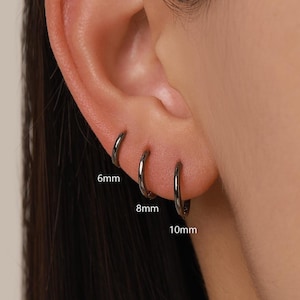 May include: Three black hoop earrings in sizes 6mm, 8mm, and 10mm. The earrings are shown on an ear, highlighting their different diameters. The earrings have a simple, minimalist design.