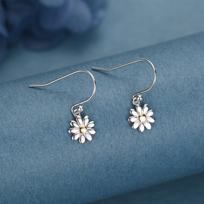 May include: A pair of silver daisy earrings with white petals and a gold center. The earrings have a hook design and are displayed against a blue background.