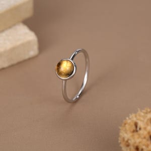 Puede incluir: Un anillo de tono plateado con una piedra cabujón redonda de color ámbar engastada en un bisel de metal liso. La banda es delgada y pulida, creando una silueta circular simple que resalta la gema de color amarillo anaranjado cálido y translúcido.