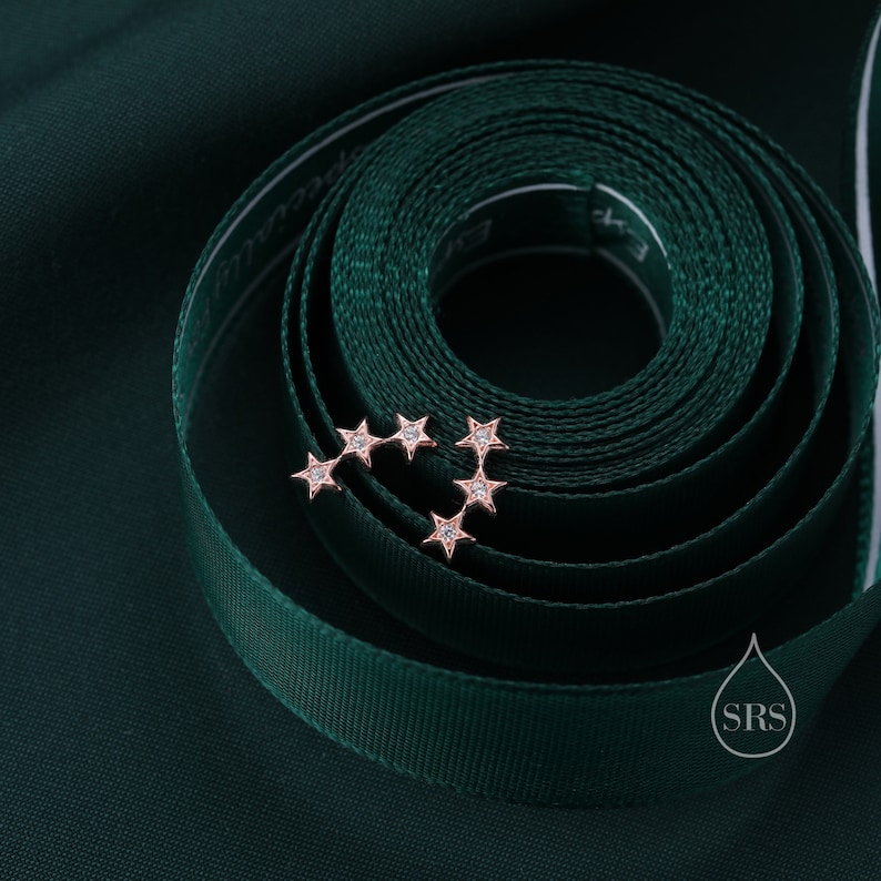 May include: A pair of rose gold star-shaped earrings with small clear gemstones set in each point of the star. The earrings are on a dark green background.