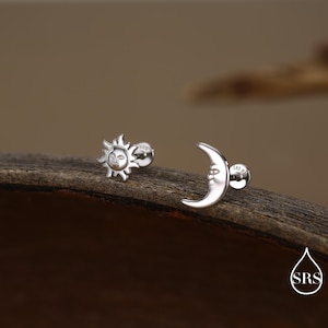 May include: Two silver stud earrings, one shaped like a sun with a face and the other a crescent moon with a face. The earrings are on a wooden surface. The sun earring has a diameter of approximately 0.3 inches.