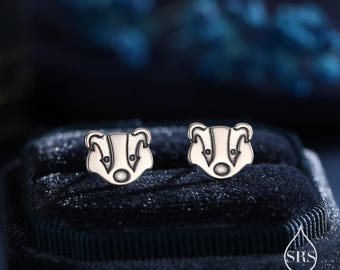 May include: A pair of silver-colored stud earrings. Each earring features a detailed badger design. The earrings are displayed on a dark blue velvet surface, with a blurred background.