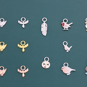 Peut inclure: Une collection de petits charms en forme d'oiseaux en argent, or et or rose. Les charms comprennent une plume, un rouge-gorge, un pingouin et d'autres motifs d'oiseaux. Chaque charm a une petite boucle.