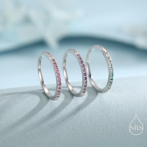 Op de afbeelding: Drie zilveren ringen met gekleurde edelstenen. Eén ring heeft roze stenen, de tweede paarse stenen en de derde groene stenen. De ringen worden gepresenteerd op een lichtblauwe ondergrond.