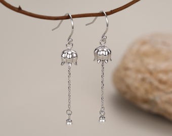 May include: A pair of silver-toned drop earrings featuring a small, bell-shaped flower charm suspended from a delicate chain. Each earring includes a curved hook wire for attachment and a tiny, smooth spherical bead at the end of the chain.
